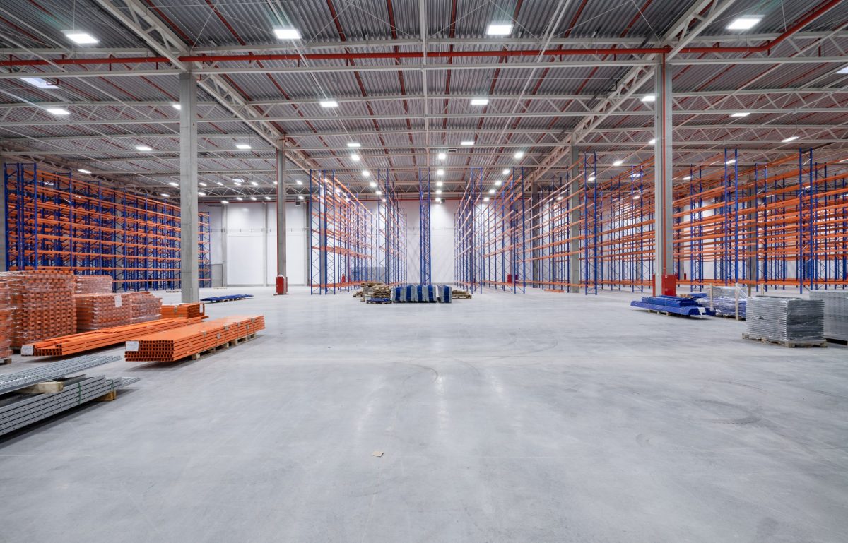 A large, well-lit, empty warehouse with polished concrete floors and stacks of grey and orange pallets in the midground.