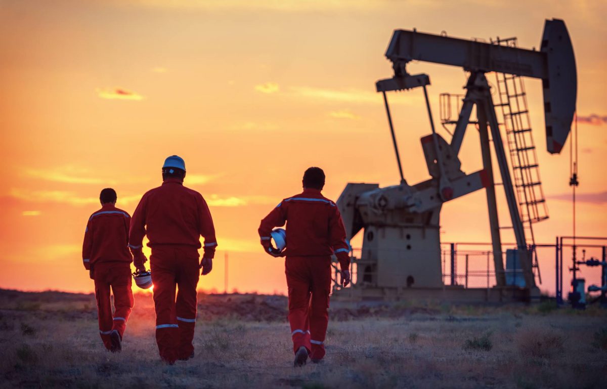 Three men wearing protective gear, walking toward an oilfield. The sun is rising and a large machine sits in the background.
