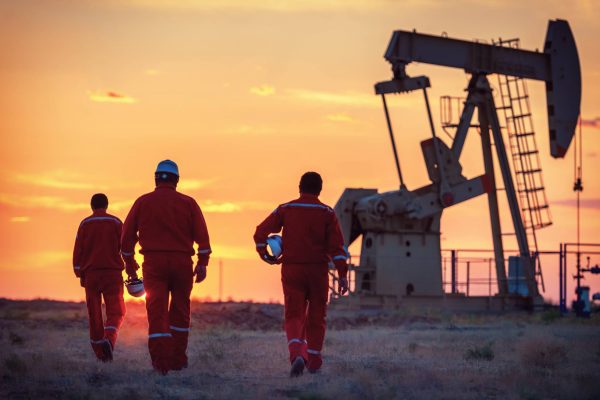 Three men wearing protective gear, walking toward an oilfield. The sun is rising and a large machine sits in the background.