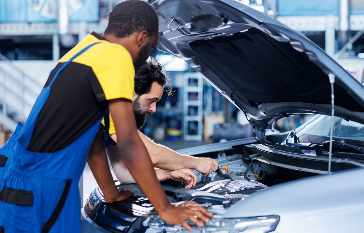 Two male auto repair technicians work together to diagnose and treat the engine of gray sedan with its hood open.