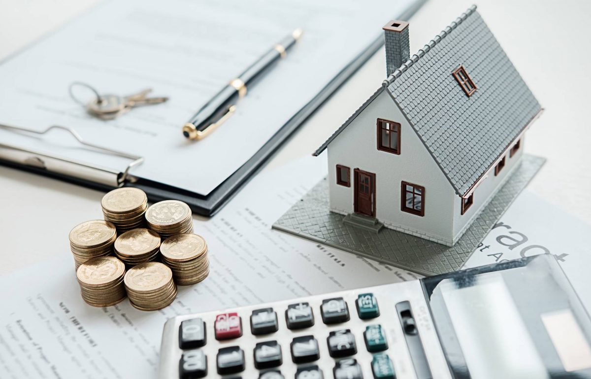 A close-up of a plastic house model on top of a real estate contract with coin money stacked up and a calculator on the desk.