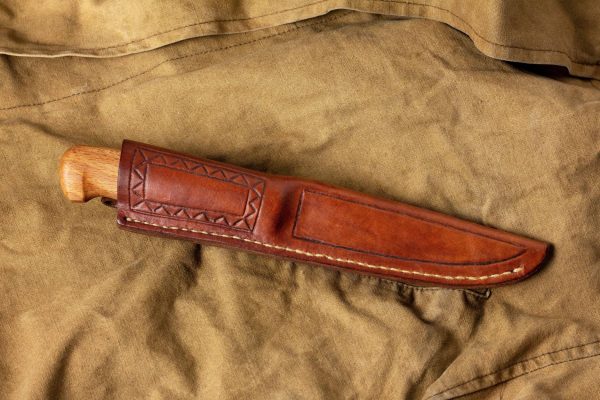 A knife with a fixed blade in a patterned brown leather sheath on a wrinkled tan cloth background.