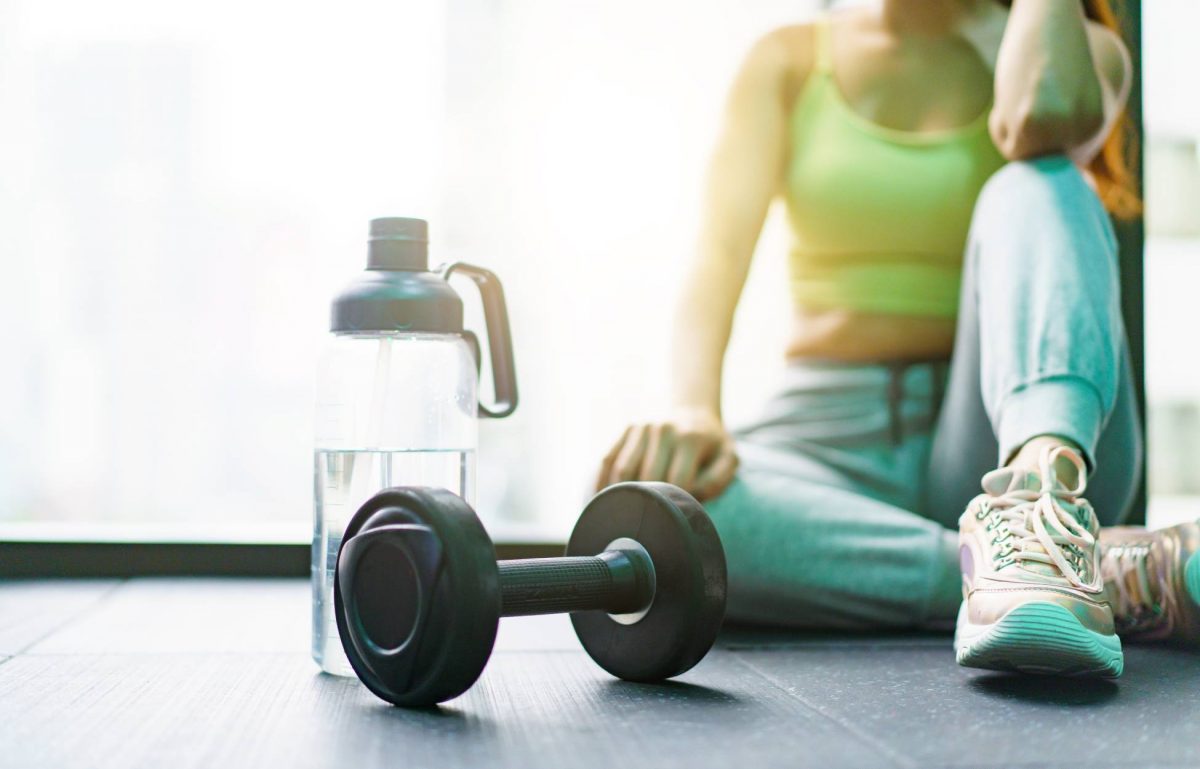 A woman in a tank top, sweatpants, and sneakers sits on the floor. A water bottle and a dumbbell are nearby.