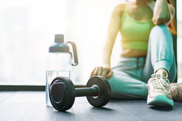 A woman in a tank top, sweatpants, and sneakers sits on the floor. A water bottle and a dumbbell are nearby.