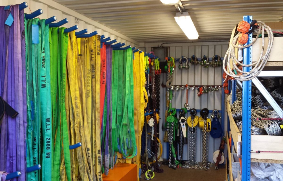A warehouse with rigging equipment hung on hooks. The equipment includes ropes in various colors and chains with hooks.