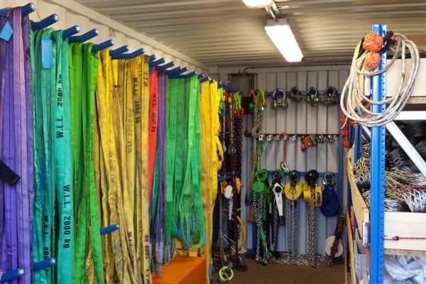 A warehouse with rigging equipment hung on hooks. The equipment includes ropes in various colors and chains with hooks.