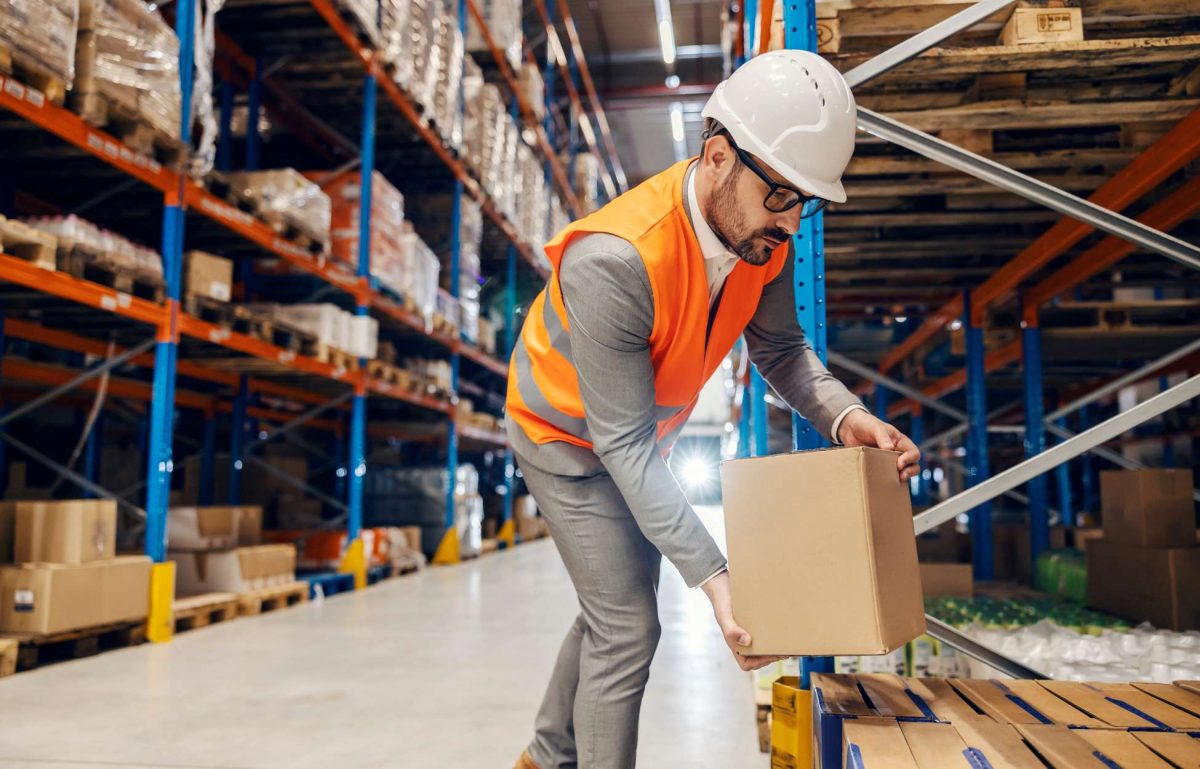 A warehouse worker wearing a hard hat and safety vest carefully placing a box onto a pallet situated on a shelving rack.