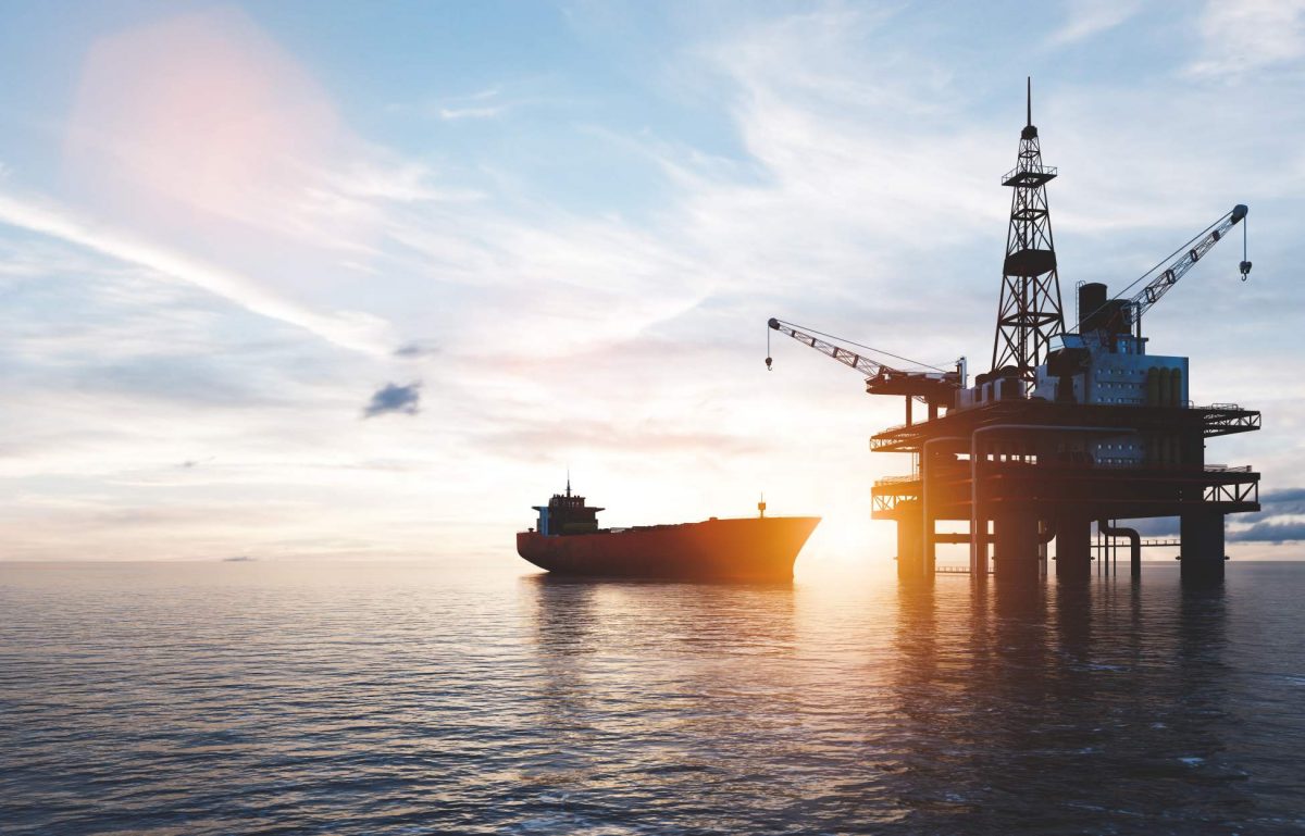 A large tanker appears behind an offshore drilling rig in the ocean. The sun is setting in the background.