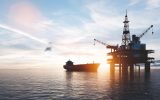 A large tanker appears behind an offshore drilling rig in the ocean. The sun is setting in the background.