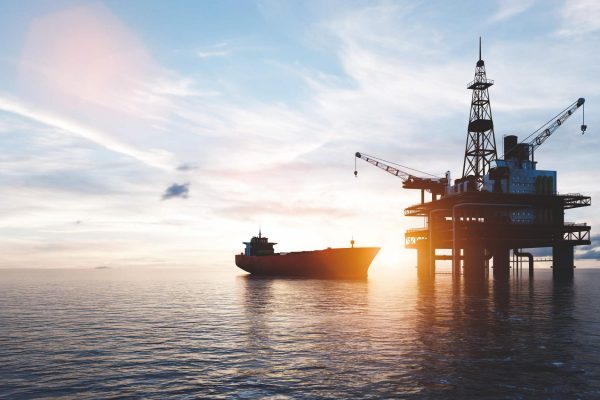 A large tanker appears behind an offshore drilling rig in the ocean. The sun is setting in the background.