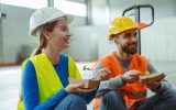 A male and female construction worker sitting next to each other as they smile and eat lunch together.