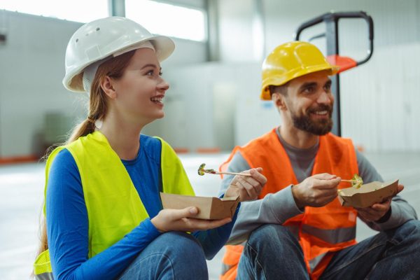 A male and female construction worker sitting next to each other as they smile and eat lunch together.