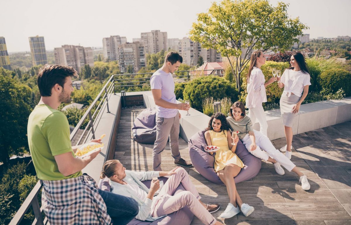 Seven young people are hanging out on a rooftop terrace, talking and smiling. They're drinking and eating.