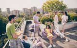 Seven young people are hanging out on a rooftop terrace, talking and smiling. They're drinking and eating.