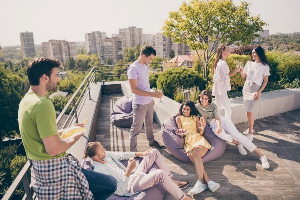 Seven young people are hanging out on a rooftop terrace, talking and smiling. They're drinking and eating.