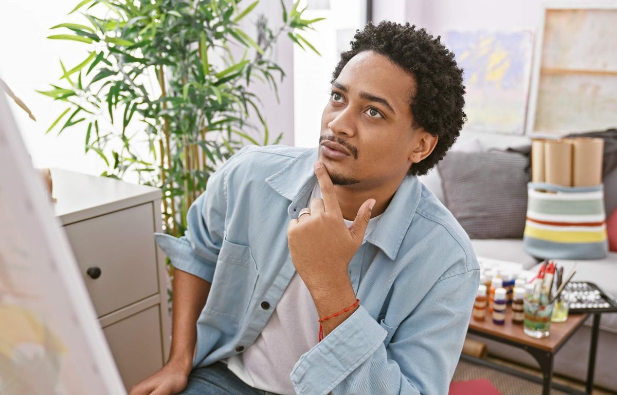 A male with curly hair in a jean jacket ponders while looking at art in an art studio or apartment setting.