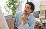 A male with curly hair in a jean jacket ponders while looking at art in an art studio or apartment setting.