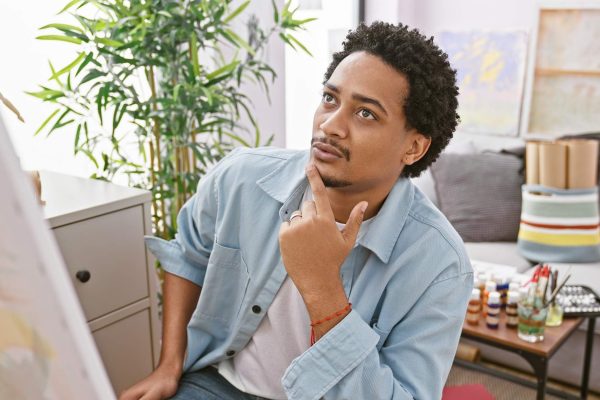 A male with curly hair in a jean jacket ponders while looking at art in an art studio or apartment setting.