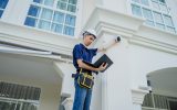 A male technician in a white hard hat sets up a CCTV camera on the exterior of a residential building.