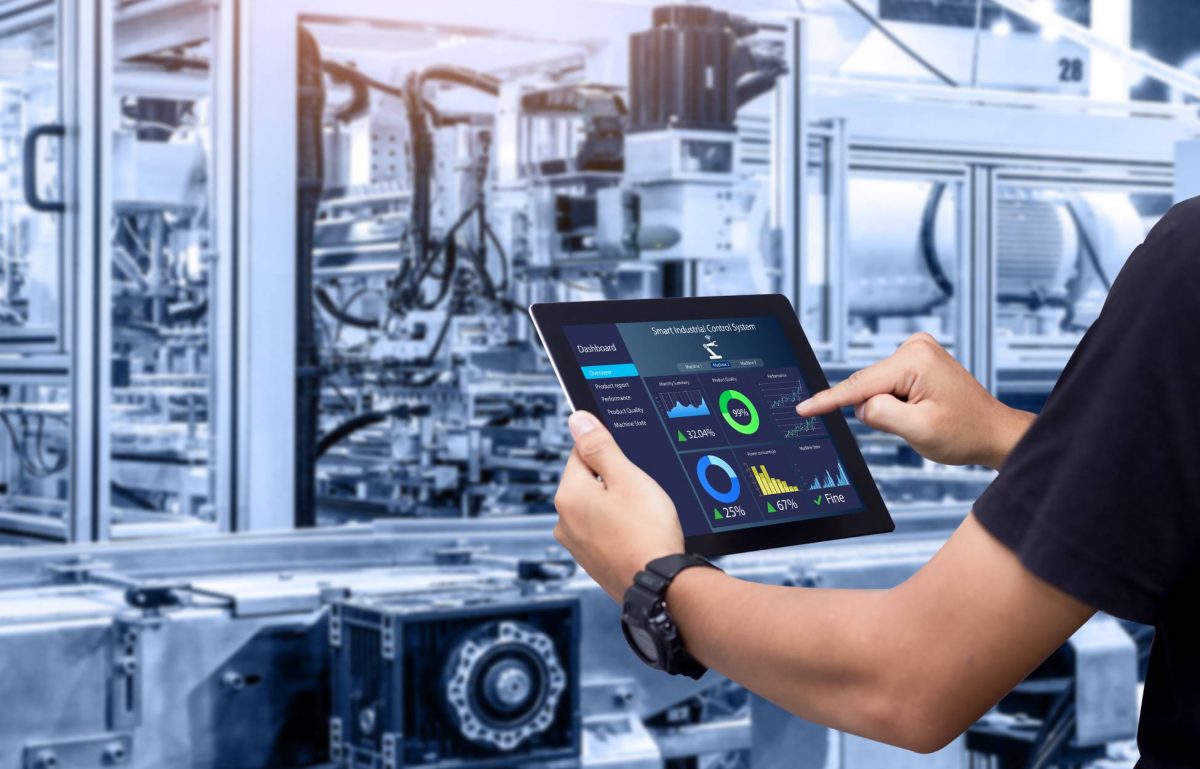 An employee in a black shirt holds a smart tablet with various controls. A blurred machine sits in the background.