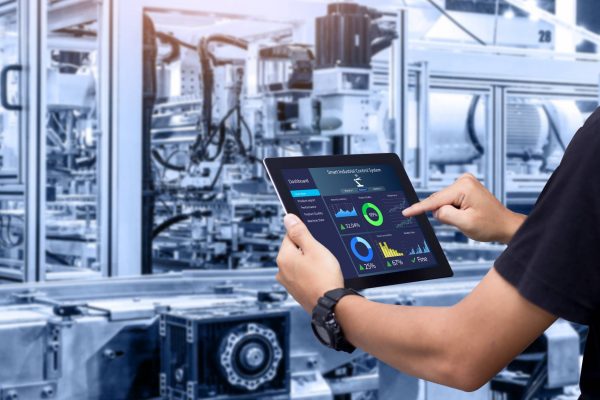 An employee in a black shirt holds a smart tablet with various controls. A blurred machine sits in the background.