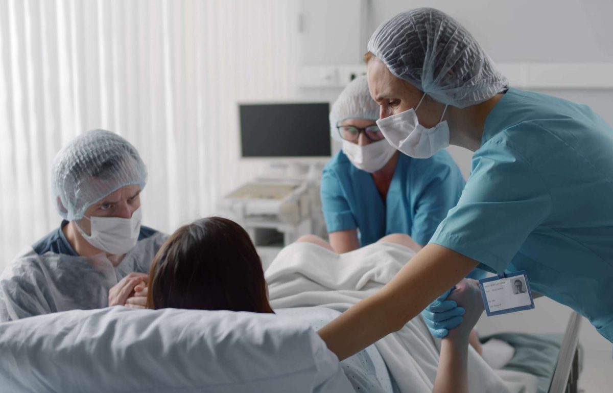 A woman is in a hospital bed giving birth. A doctor, a nurse, and her husband surround her for support.