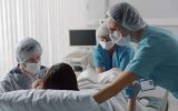 A woman is in a hospital bed giving birth. A doctor, a nurse, and her husband surround her for support.