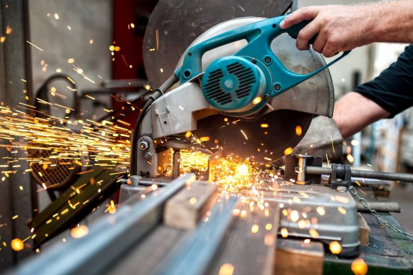 A person wearing a black shirt is using a teal saw to cut through a piece of metal. Sparks fly around the saw.