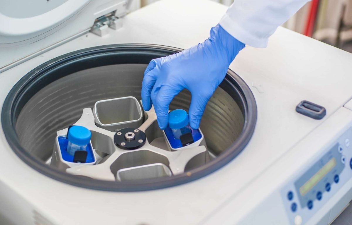 A lab tech placing a sample case into a centrifuge. They're wearing blue latex gloves and a white lab coat.