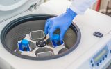 A lab tech placing a sample case into a centrifuge. They're wearing blue latex gloves and a white lab coat.