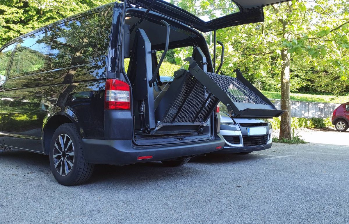 A black wheelchair-accessible vehicle sits in a parking lot. The trunk is open, and a ramp is unfolding.