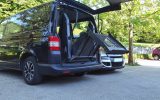 A black wheelchair-accessible vehicle sits in a parking lot. The trunk is open, and a ramp is unfolding.