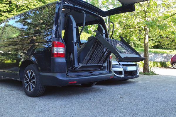 A black wheelchair-accessible vehicle sits in a parking lot. The trunk is open, and a ramp is unfolding.