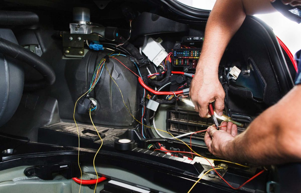 A lot of wires from within a car are exposed. An electrician holds a wire stripper against one of the wires.