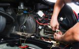 A lot of wires from within a car are exposed. An electrician holds a wire stripper against one of the wires.