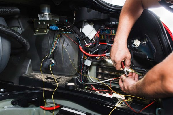 A lot of wires from within a car are exposed. An electrician holds a wire stripper against one of the wires.