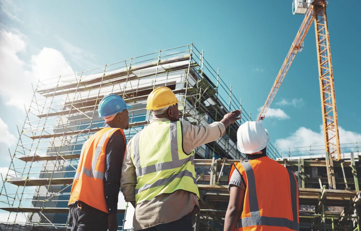 Three workers in safety vests are overlooking the construction of a large building. One is pointing to a large crane.