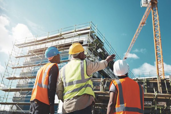 Three workers in safety vests are overlooking the construction of a large building. One is pointing to a large crane.