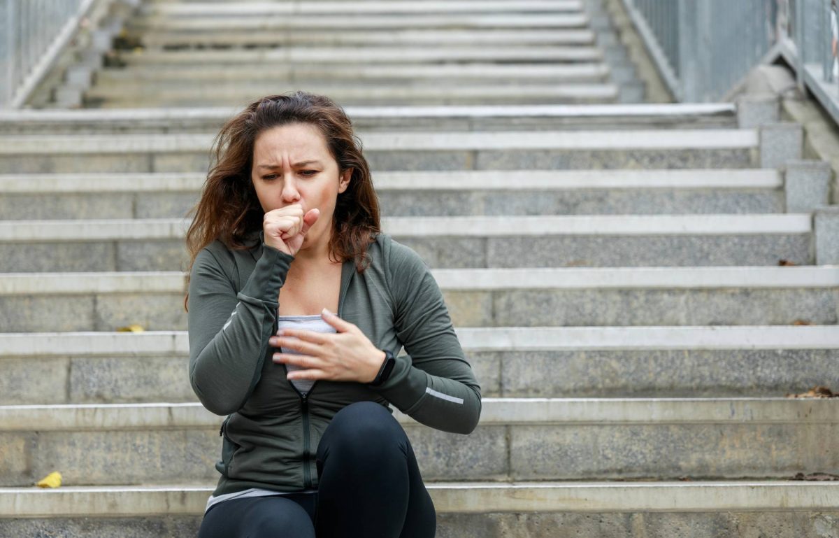 A woman sitting on an outdoor staircase and wearing athletic clothing. She is coughing into her hand.