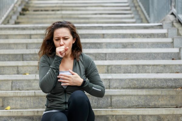 A woman sitting on an outdoor staircase and wearing athletic clothing. She is coughing into her hand.