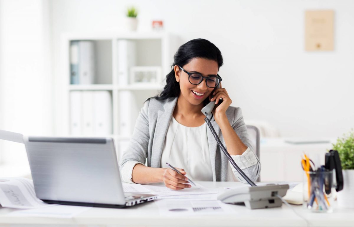 A young woman wearing a white blazer and blouse smiles as she talks on a phone at a desk. An open laptop sits on the desk.