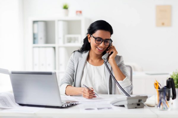 A young woman wearing a white blazer and blouse smiles as she talks on a phone at a desk. An open laptop sits on the desk.