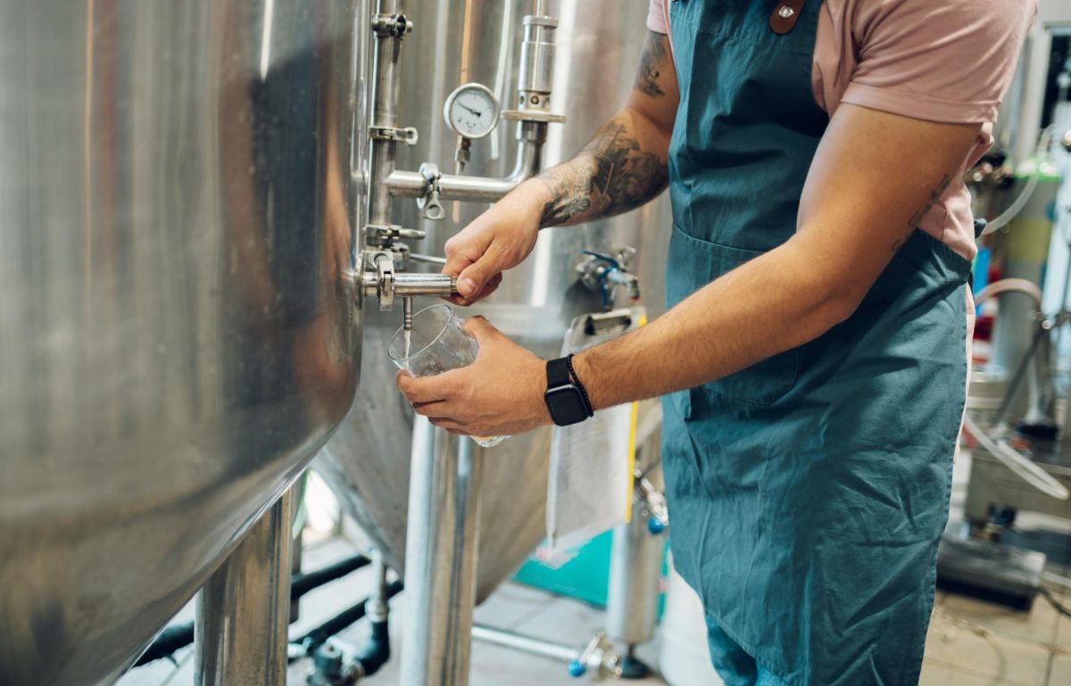 A brewer wearing a green apron and holding a glass next to a stainless steel tank. He pours a sample of beer into the glass.