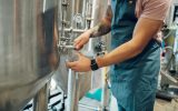 A brewer wearing a green apron and holding a glass next to a stainless steel tank. He pours a sample of beer into the glass.