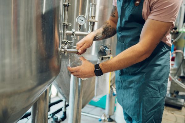 A brewer wearing a green apron and holding a glass next to a stainless steel tank. He pours a sample of beer into the glass.