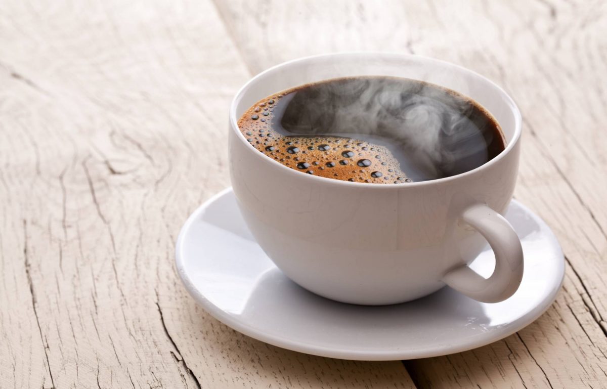 A steaming cup of brown coffee sits in a small dish on a wooden table. The table consists of white planks.