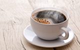 A steaming cup of brown coffee sits in a small dish on a wooden table. The table consists of white planks.