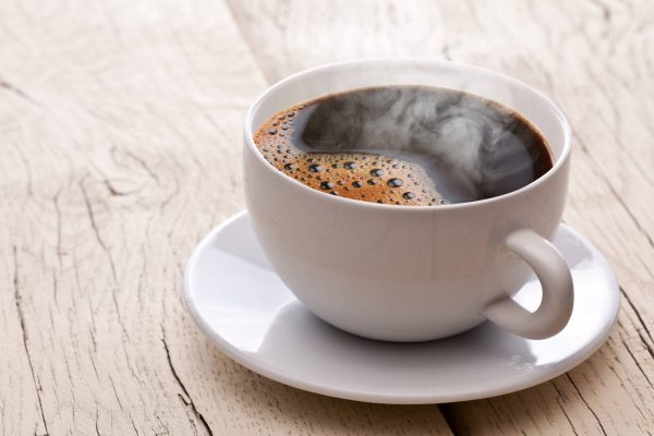 A steaming cup of brown coffee sits in a small dish on a wooden table. The table consists of white planks.