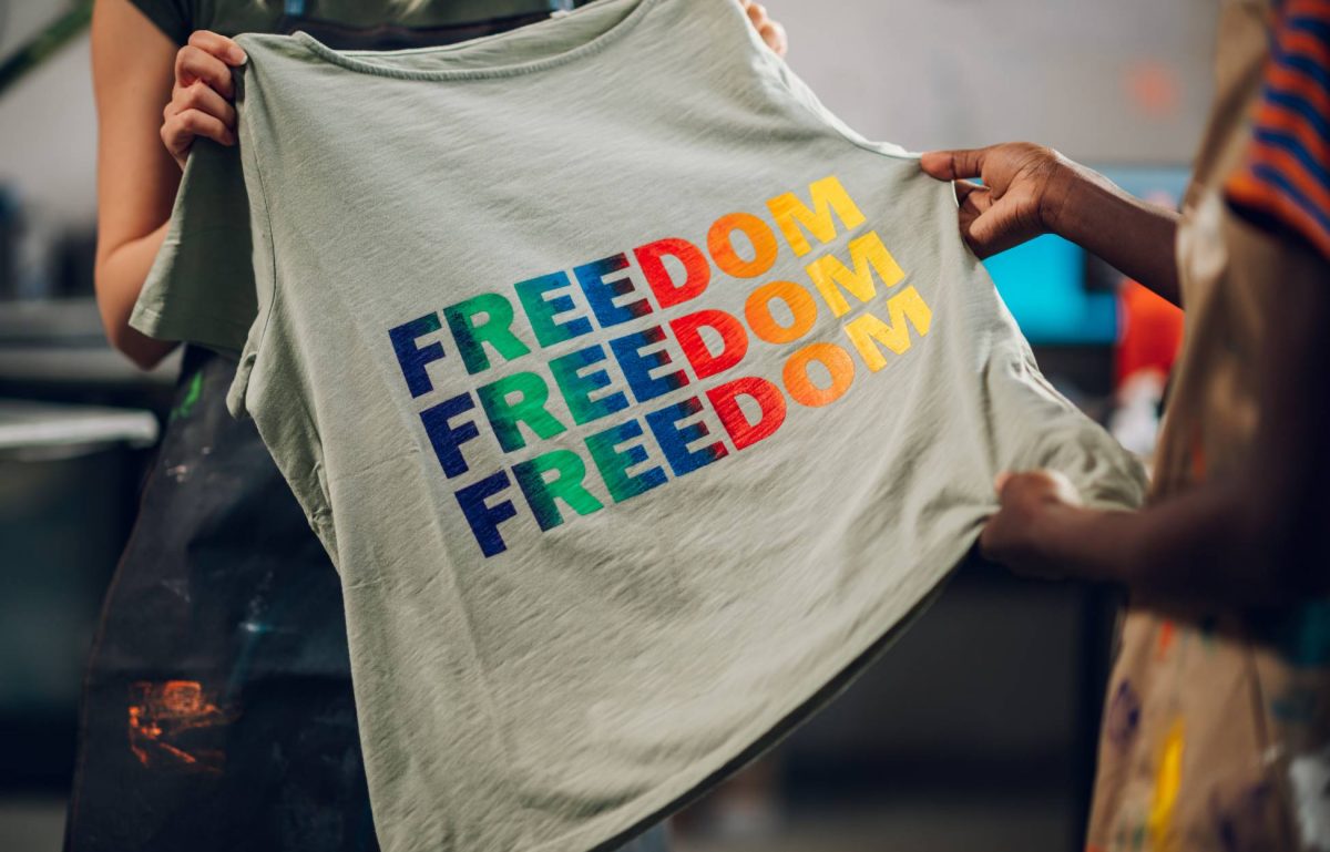 Two textile workers holding up a grey t-shirt with the word "freedom" printed three times on the shirt in bright, rainbow colors.