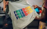 Two textile workers holding up a grey t-shirt with the word "freedom" printed three times on the shirt in bright, rainbow colors.